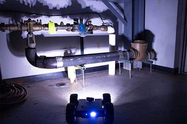 UplinkRobotics inspection crawler with 4 wheel drive driving with lights on in a dark industrial pipe space.